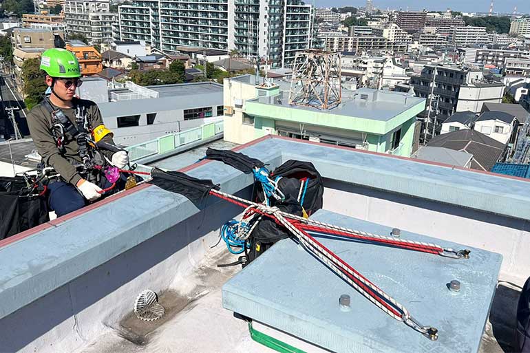 屋上のアンカーにロープを設置し、ハーネスとヘルメットを着用した作業員が降下準備をしている様子