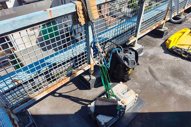 屋上に積み重ねたカウンターウエイトにスリングを接続し、フェンス越しにロープを設置して吊り元を確保している様子